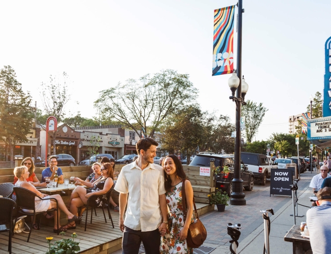 couple walking down the street holding hands but patio tables set out on the street