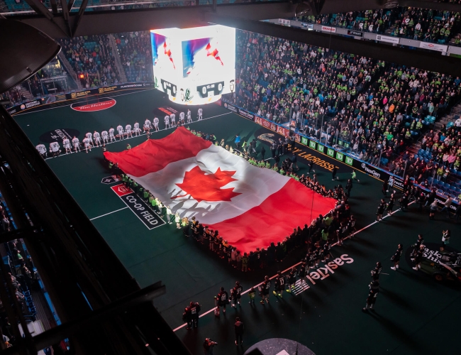 A football stadium fulled with fans. There is a demonstration on the field were individuals are holding a huge Canadian flag