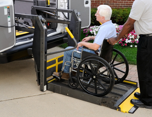 An elderly man being assisted on to a bus with a chair lift. 