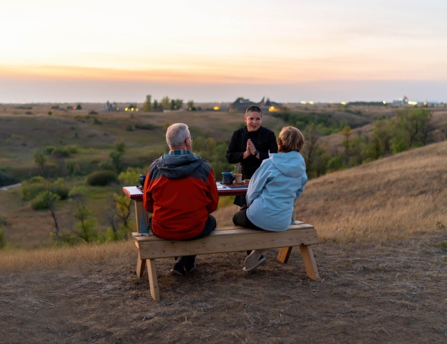 Couple sitting at an outdoor patio table set with food and drinks, while a host speaks to them