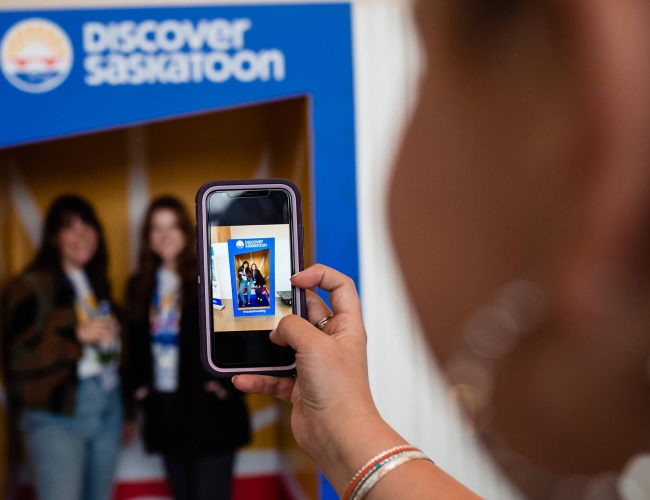 Person taking a photo of 2 women in front of a Discover Saskatoon backdrop