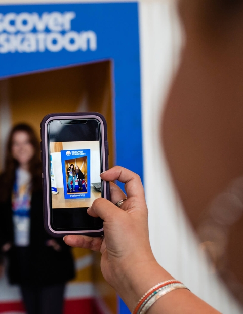 Person taking a photo of 2 women in front of a Discover Saskatoon backdrop